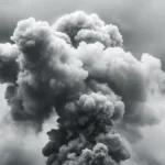 Dark gray smoke cloud rising against a light clouded sky, showing turbulent movement.
