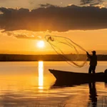 Fisherman casting a net on a tranquil lake during sunset with reeds nearby.
