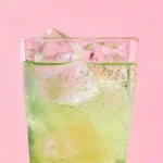 Green fizzy drink in a tall glass with ice on pink background.