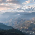 Snow-capped mountains with pastel sky over rolling hills and winding roads.