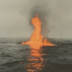 Large flame erupting from the ocean during a volcanic eruption with smoke and dark waves.