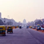 Street scene in New Delhi with tuk-tuks, cars, and people bustling.