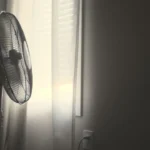 White wall fan running near white window blinds in hot bedroom.