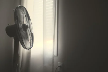 White wall fan running near white window blinds in hot bedroom.