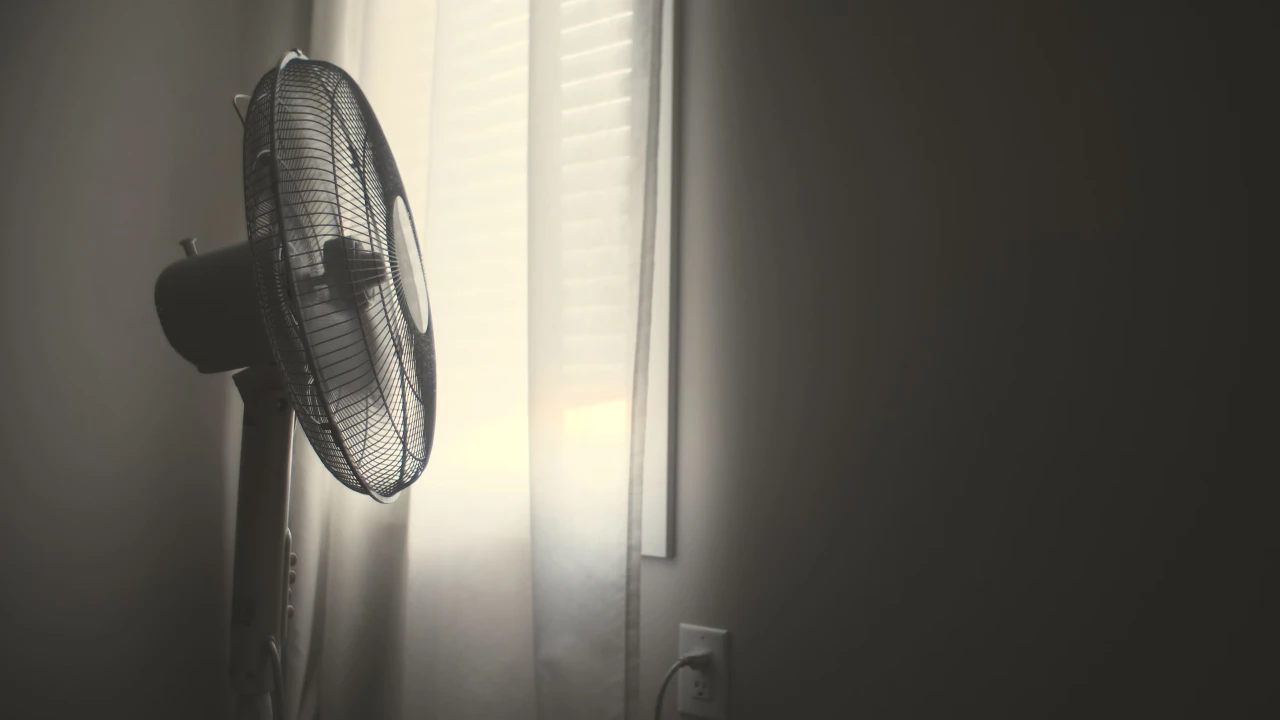 White wall fan running near white window blinds in hot bedroom.